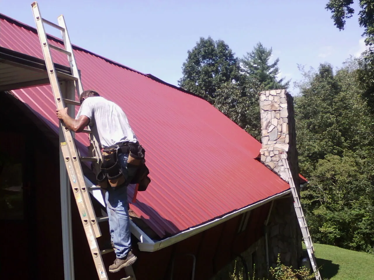 Roofer on a ladder beside a red metal roof and stone chimney — Soffit & Fascia Repair by Roofing Brothers in Blackstone